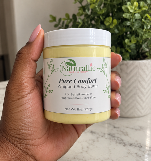 A hand holding a jar of Naturallie Pure Comfort Whipped Body Butter, with a potted green plant in the background.