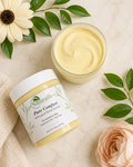 Naturalie Pure Comfort Whipped Body Butter in a clear jar, shown next to an open jar of the pale yellow whipped balm on a light marble surface with green leaves, a daisy, and a peach rose.