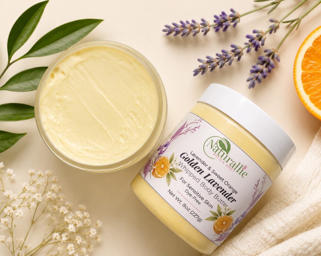 A jar of Naturlie Golden Lavender whipped body butter next to an open jar showing the pale yellow cream, styled with lavender sprigs, baby's breath, green leaves, and an orange slice on a beige fabric background.